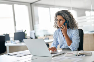 woman in blue shirt talking on the phone and typing on laptop